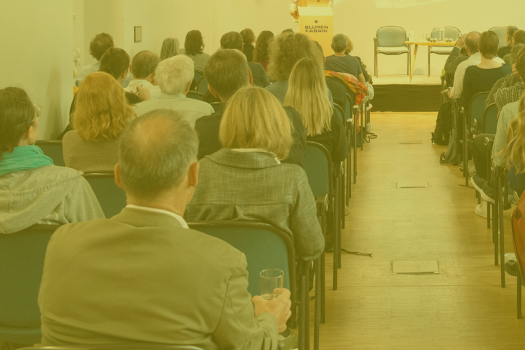 Publikum von hinten, das in Richtung Bühne schaut, auf dem ein Mann hinter einem Pult zu sehen ist. Sein Oberkörper ist vom Bildrand abgeschnitten