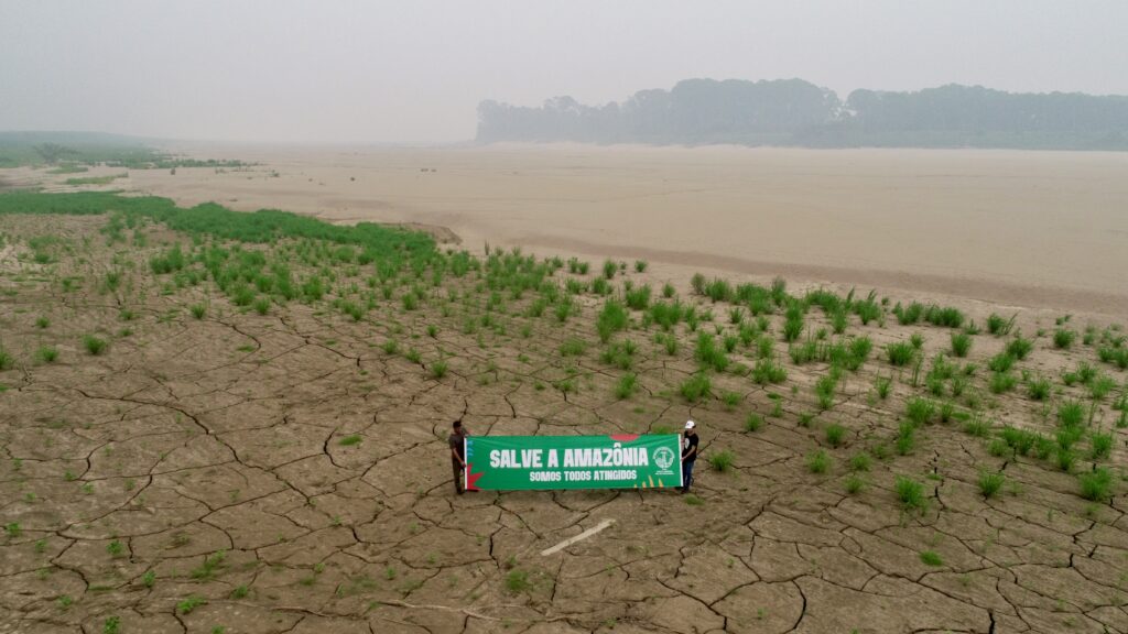 Bilaterale Klimaprojekte: Zwei Personen halten ein grünes Protestschild, im Hintergrund ist das Wasser des Staudammbeckens zu sehen.