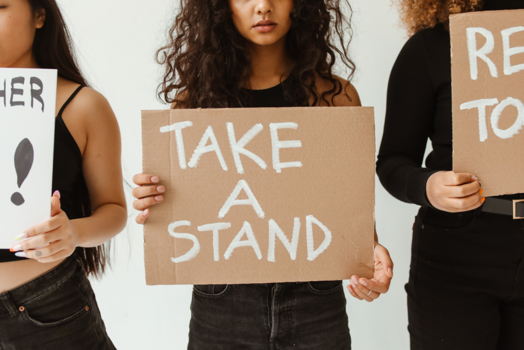 Personen stehen nebeneinander und halten Schilder hoch. Im Zentrum steht eine Person mit einem Schild, auf dem "Take a stand" steht.